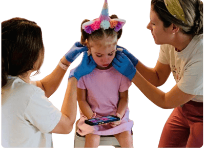 Young girl with a unicorn headband getting both ears pierced at the same time by two professionals wearing gloves, with flower illustrations in the background. Simultaneous ear piercing service at Peekaboo Piercing in Tampa.
